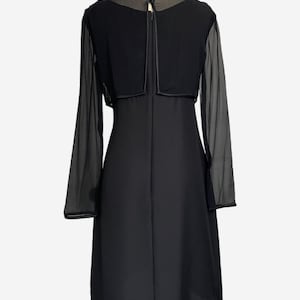 K&ouml;nnte beinhalten: Ein schwarzes Kleid mit langen, transparenten &Auml;rmeln und einem passenden Bolero-Oberteil. Das Kleid hat ein schlichtes, elegantes Design mit einem figurbetonten Oberteil und einem knielangen Rock. Der Bolero hat einen runden Ausschnitt und einen kleinen Knopfverschluss am R&uuml;cken.