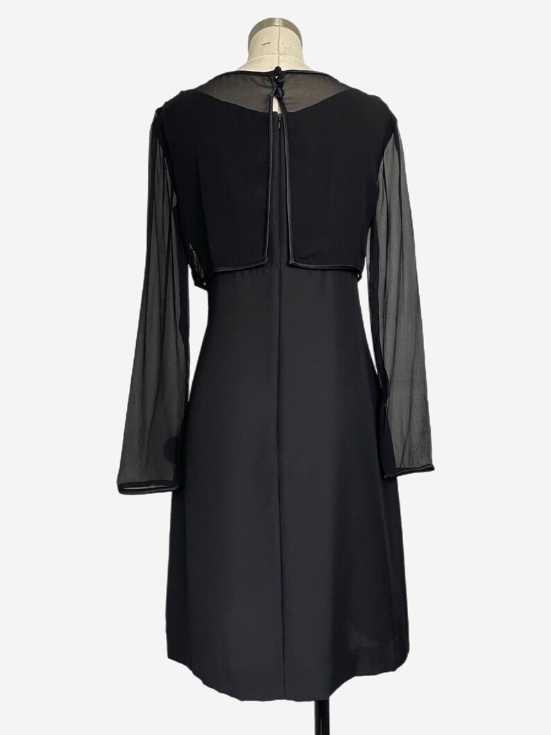 K&ouml;nnte beinhalten: Ein schwarzes Kleid mit langen, transparenten &Auml;rmeln und einem passenden Bolero-Oberteil. Das Kleid hat ein schlichtes, elegantes Design mit einem figurbetonten Oberteil und einem knielangen Rock. Der Bolero hat einen runden Ausschnitt und einen kleinen Knopfverschluss am R&uuml;cken.