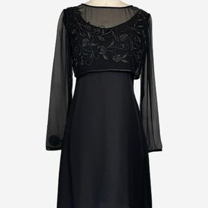 K&ouml;nnte beinhalten: Ein schwarzes Kleid mit einem transparenten &Uuml;berzug mit floraler Stickerei. Das Kleid hat lange &Auml;rmel mit Kn&ouml;pfen an den Manschetten. Das Oberteil ist figurbetont und der Rock f&auml;llt ab der Taille aus. Das Kleid wird auf einer Schaufensterpuppe pr&auml;sentiert.