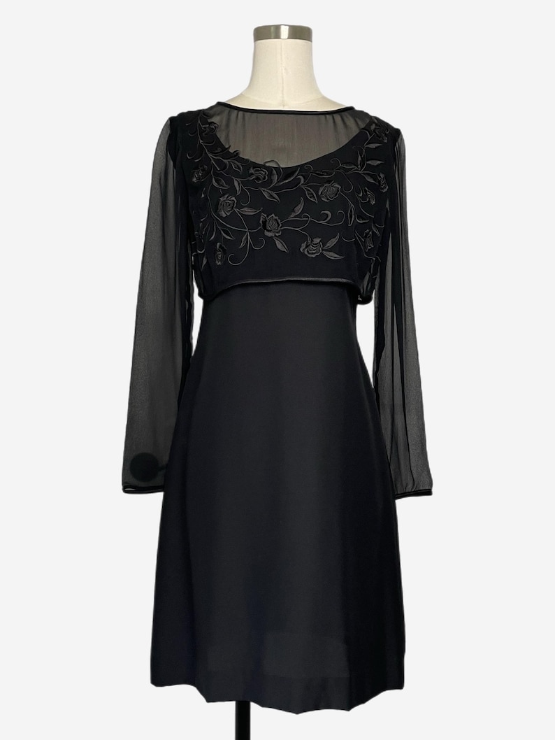 K&ouml;nnte beinhalten: Ein schwarzes Kleid mit einem transparenten &Uuml;berzug mit floraler Stickerei. Das Kleid hat lange &Auml;rmel mit Kn&ouml;pfen an den Manschetten. Das Oberteil ist figurbetont und der Rock f&auml;llt ab der Taille aus. Das Kleid wird auf einer Schaufensterpuppe pr&auml;sentiert.