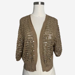 May include: A tan, open-front cardigan with short sleeves. The knitwear features a loose, open weave and is embellished with small, round sequins. The cardigan is displayed on a mannequin against a plain white background.