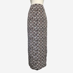 May include: A long, patterned skirt with a repeating square design in shades of brown, black, and white. The skirt is displayed on a mannequin, showcasing its full length and fitted silhouette. The fabric appears to be lightweight.