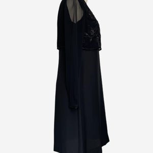 K&ouml;nnte beinhalten: Schwarzes Kleid mit transparentem Overlay an Schultern und &Auml;rmeln. Das Oberteil hat ein gesticktes Blumenmuster. Das Kleid wird auf einer Schaufensterpuppe vor wei&szlig;em Hintergrund pr&auml;sentiert.