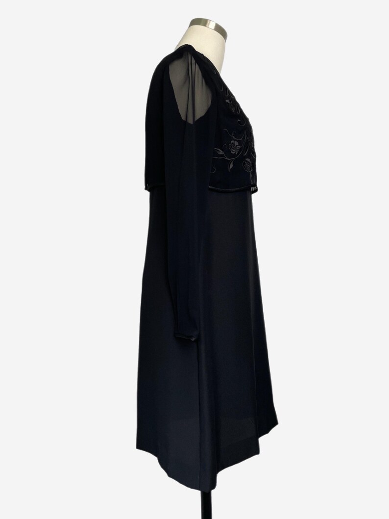 K&ouml;nnte beinhalten: Schwarzes Kleid mit transparentem Overlay an Schultern und &Auml;rmeln. Das Oberteil hat ein gesticktes Blumenmuster. Das Kleid wird auf einer Schaufensterpuppe vor wei&szlig;em Hintergrund pr&auml;sentiert.
