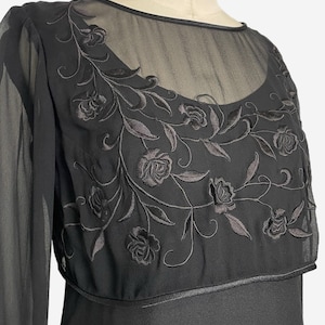 K&ouml;nnte beinhalten: Schwarzes Kleid mit langen &Auml;rmeln und einem transparenten Overlay. Das Overlay weist gestickte florale Details auf Brust und Schultern auf. Das Kleid hat einen runden Ausschnitt und ein figurbetontes Oberteil.