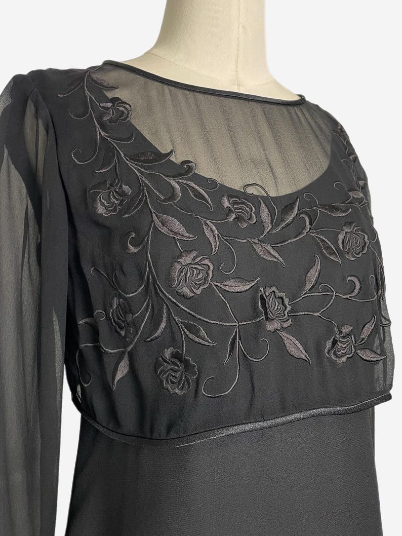 K&ouml;nnte beinhalten: Schwarzes Kleid mit langen &Auml;rmeln und einem transparenten Overlay. Das Overlay weist gestickte florale Details auf Brust und Schultern auf. Das Kleid hat einen runden Ausschnitt und ein figurbetontes Oberteil.