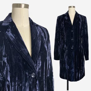 May include: A navy blue velvet coat with a classic lapel collar and three buttons. The long coat is displayed on a mannequin and has a slightly textured appearance. The coat is a solid color and has a tailored fit.