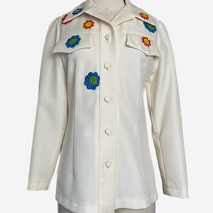 May include: Cream-colored button-down shirt with colorful floral appliqués. The shirt features two chest pockets and long sleeves. The flowers are in shades of red, blue, yellow, and green, adding a pop of color to the vintage garment.