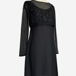K&ouml;nnte beinhalten: Ein schwarzes Kleid mit einem transparenten &Uuml;berzug und langen &Auml;rmeln. Der &Uuml;berzug hat einen runden Ausschnitt und ist mit floralen Stickereien verziert. Das Kleid wird auf einer Schaufensterpuppe pr&auml;sentiert.