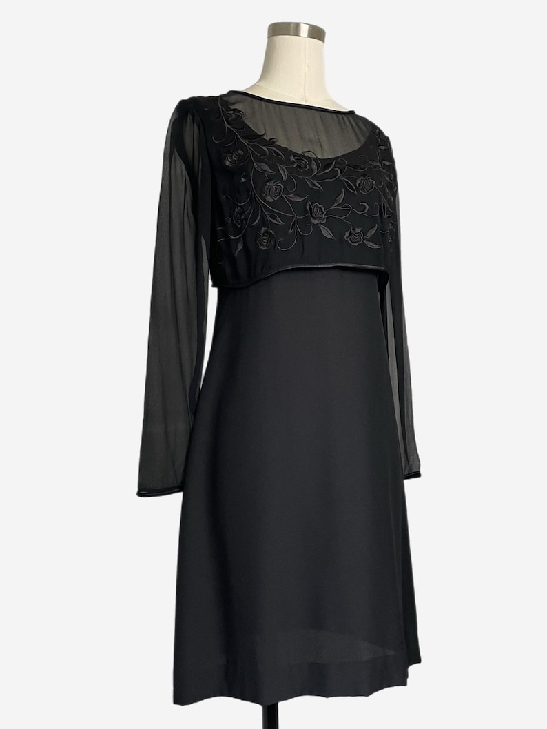 K&ouml;nnte beinhalten: Ein schwarzes Kleid mit einem transparenten &Uuml;berzug und langen &Auml;rmeln. Der &Uuml;berzug hat einen runden Ausschnitt und ist mit floralen Stickereien verziert. Das Kleid wird auf einer Schaufensterpuppe pr&auml;sentiert.