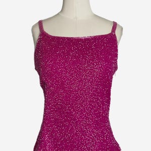 May include: A magenta-colored beaded tank top with thin straps. The top is displayed on a mannequin. The fabric is covered in small, shiny beads, creating a textured appearance. The neckline is square, and the hemline has a slight ruffle.
