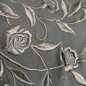 K&ouml;nnte beinhalten: Nahaufnahme eines dunkelgrauen Stoffes mit gestickten Blumenmustern. Die Stickerei zeigt detaillierte Rosen und Bl&auml;tter in einem helleren Grau, wodurch ein strukturiertes, elegantes Design entsteht. Der Stoff scheint ein Kleid oder ein anderes Kleidungsst&uuml;ck zu sein.