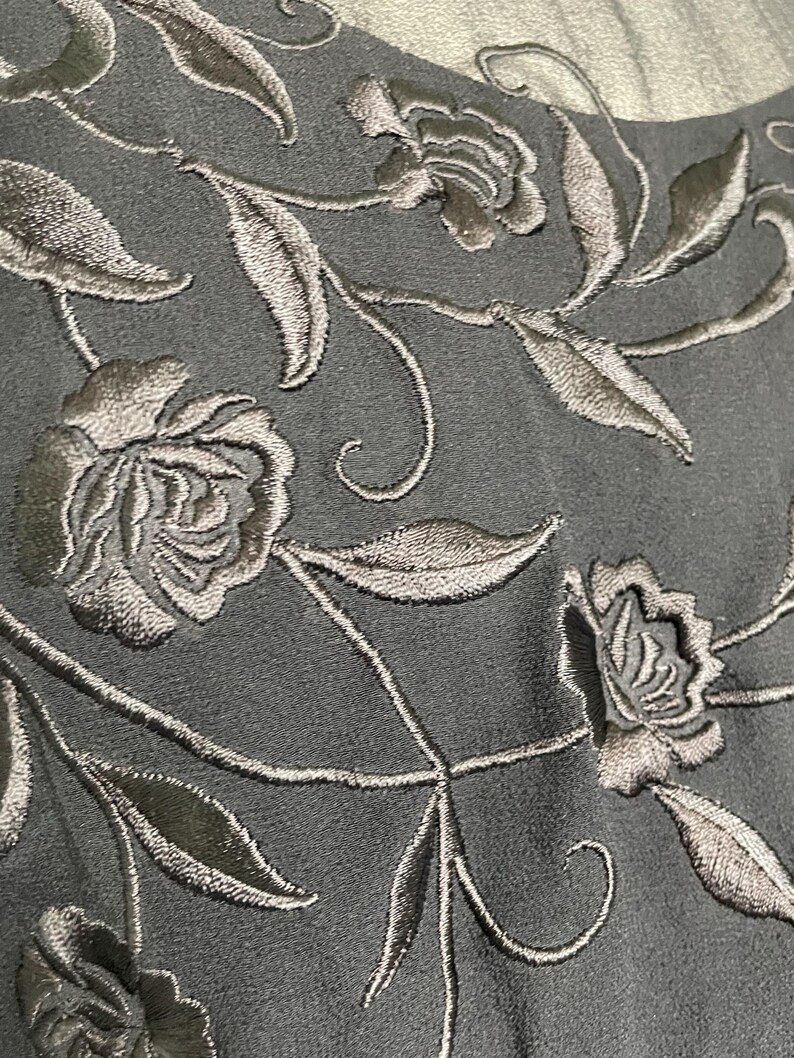 K&ouml;nnte beinhalten: Nahaufnahme eines dunkelgrauen Stoffes mit gestickten Blumenmustern. Die Stickerei zeigt detaillierte Rosen und Bl&auml;tter in einem helleren Grau, wodurch ein strukturiertes, elegantes Design entsteht. Der Stoff scheint ein Kleid oder ein anderes Kleidungsst&uuml;ck zu sein.