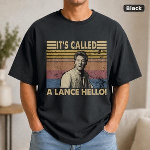 May include: A black short-sleeve t-shirt featuring a vintage-style graphic with horizontal stripes in muted colors. The design includes a portrait of a person and the text "IT'S CALLED A LANCE HELLO!" printed in a bold, retro-inspired font.