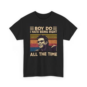 May include: Black t-shirt featuring a smiling man wearing sunglasses, with the text "BOY DO I HATE BEING RIGHT ALL THE TIME" in a retro design. The design includes horizontal stripes in shades of brown, red, and blue.