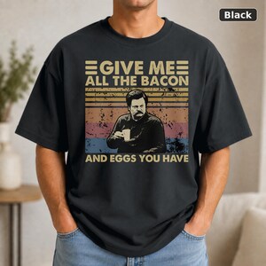 May include: A black t-shirt featuring a graphic print of a man with a mustache holding a cup. The text reads GIVE ME ALL THE BACON AND EGGS YOU HAVE. The design includes a distressed, horizontal striped background in shades of orange, yellow, and blue.