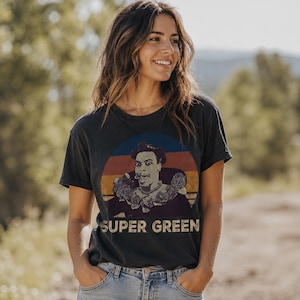 May include: A charcoal gray short-sleeve t-shirt featuring a circular graphic with a retro sunset background. The print depicts a character with a high hairstyle and a ruffled collar, with the text SUPER GREEN printed in a bold, distressed font below.