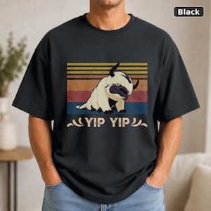 May include: A black t-shirt featuring a graphic of a cream-colored, furry creature with horns. The design includes a sunset-colored striped background and the text YIP YIP in a distressed font. The shirt has short sleeves and a crew neckline.
