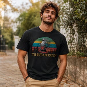 May include: A black short-sleeve t-shirt featuring a graphic of a knight helmet with a red emblem, set against a circular sunset background with horizontal stripes in yellow, orange, and teal. The text 'TIS BUT A SCRATCH' is printed in yellow.