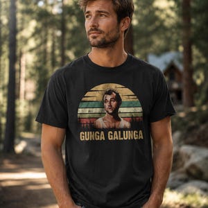 May include: A black short-sleeve t-shirt featuring a circular graphic with a retro sunset background, a portrait of a person in a white tank top, and the text "GUNGA GALUNGA" printed in a bold, yellow, sans-serif font below the illustration.