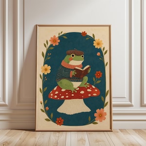 May include: A framed wall art print featuring a green frog wearing a brown beret and a blue sweater, sitting on a red and white spotted mushroom while reading an open book. The scene is enclosed in a dark blue oval border with floral accents.