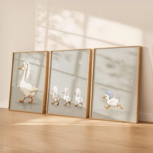 May include: Set of three framed art prints featuring whimsical illustrations of geese and ducklings. The artwork depicts a goose with a towel on its head, and ducklings wearing shower caps. The prints have a light green background and wooden frames.