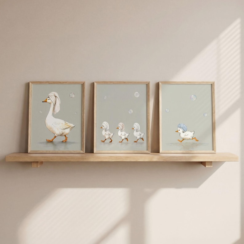 May include: Three framed art prints featuring whimsical illustrations of geese and ducklings. The first print shows a goose wearing a towel, while the others depict ducklings in bath hats. The prints are displayed on a wooden shelf against a neutral wall.