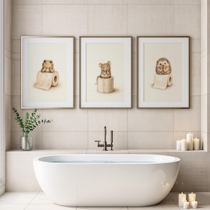 May include: Three framed wall art prints featuring illustrations of a frog, a mouse, and a hedgehog, each sitting atop a roll of toilet paper. The prints are displayed in thin gold frames against a light-coloured tiled bathroom wall above a bathtub.