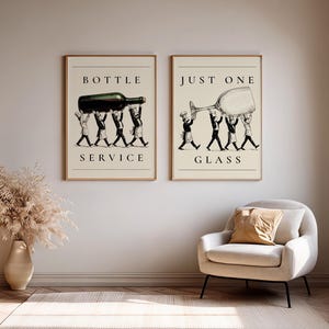 May include: Two framed wall art prints with a cream background. The left print shows four chefs carrying a dark wine bottle with the text BOTTLE SERVICE. The right print shows four chefs carrying a large wine glass with the text JUST ONE GLASS.