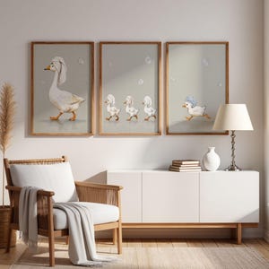 May include: Set of three framed art prints featuring watercolor illustrations of geese and ducklings. The first print shows a goose with a towel on its head, the second shows three ducklings, and the third shows a duckling running. The frames are wooden.