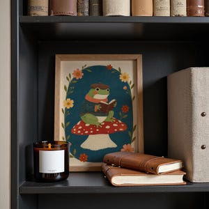May include: A framed rectangular print featuring a green frog wearing a brown beret and a striped sweater, reading a book while sitting on a red and white spotted mushroom, surrounded by a floral border on a dark blue background.