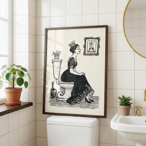 Pode incluir: Uma impressão artística a preto e branco emoldurada que mostra uma mulher com um vestido comprido e uma coroa sentada numa sanita. Uma pequena planta em vaso está sobre o autoclismo e um retrato emoldurado de um corgi com coroa.