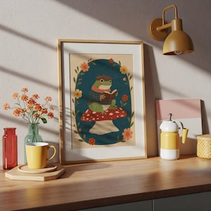 May include: A framed wall art print featuring a green frog wearing a brown beret and a blue sweater, sitting on a red and white spotted mushroom while reading a book, surrounded by an oval border of orange and yellow flowers on a dark background.