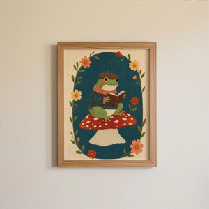 May include: A framed wall art print featuring a green frog wearing a brown acorn hat and a blue sweater, sitting on a red and white spotted mushroom while reading an open book. The illustration is set against a dark blue oval background.