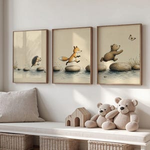 Può includere: Tre stampe da parete incorniciate con illustrazioni ad acquerello di un riccio, una volpe e un orso che saltano su pietre in un ruscello. La stampa dell'orso include una piccola farfalla. Le opere sono inserite in sottili cornici di legno.