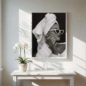 May include: Black and white framed print of a person wearing white sunglasses, a towel wrapped around their head, and a pearl necklace. The image is displayed on a white wall, with a white table and a potted orchid in the foreground.
