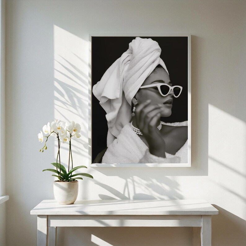 May include: Black and white framed print of a person wearing white sunglasses, a towel wrapped around their head, and a pearl necklace. The image is displayed on a white wall, with a white table and a potted orchid in the foreground.