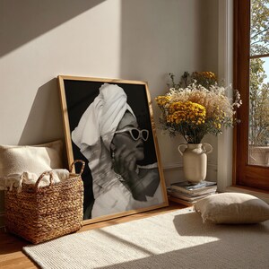 May include: A framed black and white print of a person wearing sunglasses and a towel on their head. The print is leaning against a wall. A woven basket, a vase of flowers, and a rug are in the foreground.
