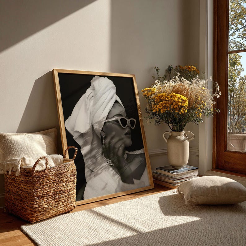 May include: A framed black and white print of a person wearing sunglasses and a towel on their head. The print is leaning against a wall. A woven basket, a vase of flowers, and a rug are in the foreground.