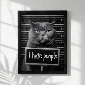 May include: A black and white framed wall art print featuring a cat in a mugshot pose against a height chart background. The cat holds a small sign with the text i hate people written in white lowercase letters. The frame is black and rectangular.