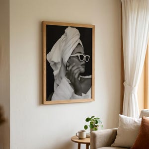 May include: Black and white framed print of a person wearing sunglasses and a towel wrapped around their head. The person is wearing a pearl necklace and earrings. The print is in a light wood frame.