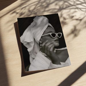 May include: Black and white photograph of a person wearing white cat-eye sunglasses, a pearl necklace, and earrings. The person is wrapped in a white towel and a white robe. The image is on a wooden surface.