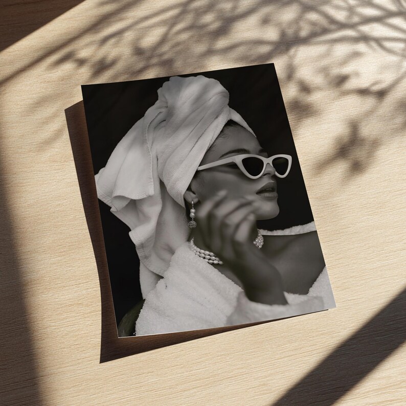 May include: Black and white photograph of a person wearing white cat-eye sunglasses, a pearl necklace, and earrings. The person is wrapped in a white towel and a white robe. The image is on a wooden surface.