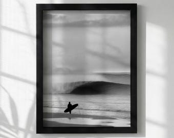Può includere: Stampa incorniciata in bianco e nero di un surfista in spiaggia, con una tavola da surf. Le onde dell'oceano sono grandi e si infrangono sullo sfondo, con un cielo nuvoloso sopra. L'immagine è racchiusa in una cornice rettangolare nera.