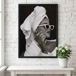 May include: Black and white framed print of a person wearing white sunglasses, a towel on their head, and a pearl necklace. The person is wearing a white robe. The print is displayed on a white table against a white brick wall.