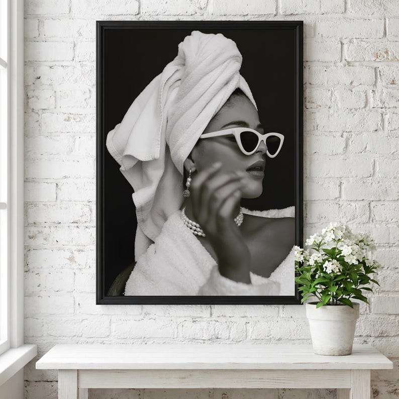 May include: Black and white framed print of a person wearing white sunglasses, a towel on their head, and a pearl necklace. The person is wearing a white robe. The print is displayed on a white table against a white brick wall.