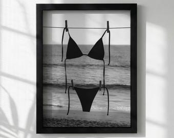 Può includere: Stampa incorniciata in bianco e nero di un bikini nero appeso a un filo con mollette di legno. Lo sfondo mostra una spiaggia e l'oceano, creando un tema estivo o vacanziero.