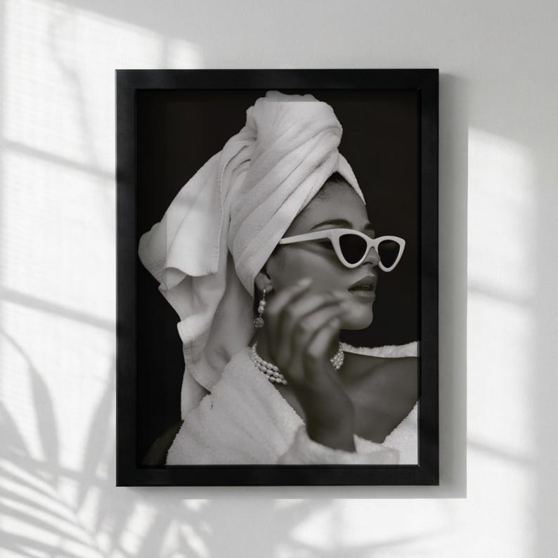 May include: Black and white framed print of a person wearing white sunglasses, a towel wrapped around their head, and a pearl necklace. The person is wearing a white robe. The image has a vintage, glamorous aesthetic.