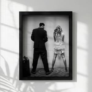 May include: A black and white framed photograph depicting two figures standing before a tiled wall. One figure wears a dark suit and stands beside a suitcase, while the other wears a light-colored dress with a ruffled skirt and high heels.