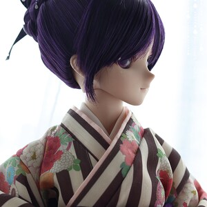 Puede incluir: Una mu&ntilde;eca con el pelo largo y morado oscuro recogido en un mo&ntilde;o con una cinta negra. La mu&ntilde;eca lleva un kimono japon&eacute;s tradicional con un estampado de rayas blancas y marrones y detalles florales. El kimono tiene una faja morada. Los ojos de la mu&ntilde;eca est&aacute;n cerrados.
